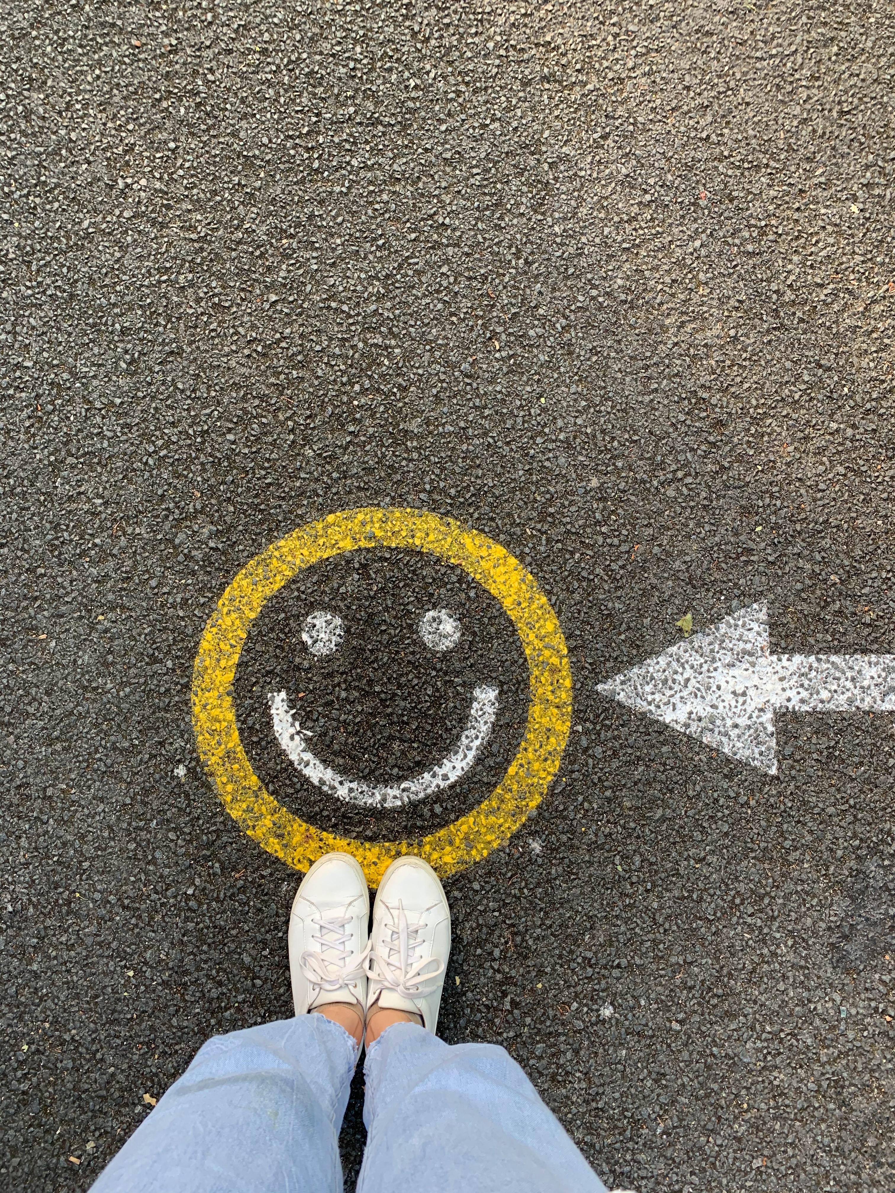 Person in white tennis shoes stands at a smiley face painted on asphalt iwth a white arrow pointing to the smiley face. 